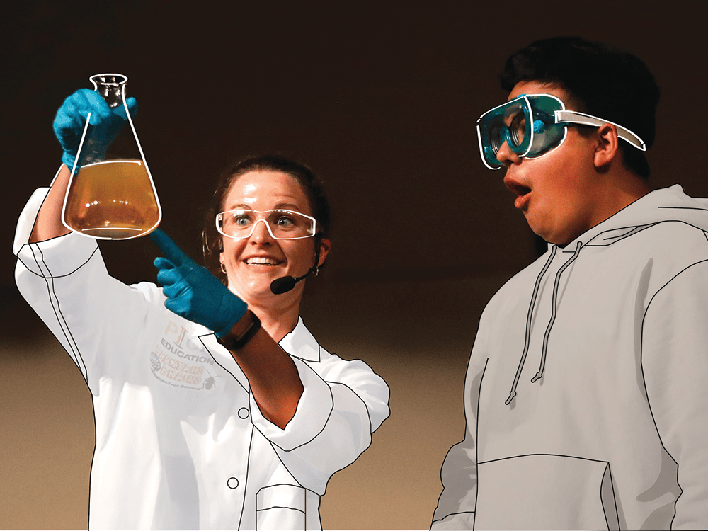 Kate Biberdorf, wearing blue gloves, a white lab coat, and safety goggles, holds up a large, conical chemistry beaker filled with orange liquid. She excitedly points at the solution to high school chemistry student Devon Zeigenbein, who wears a gray hoodie and safety goggles. He stares at the solution, mouth agape in astonishment.