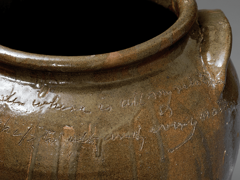 A closeup photo of a large alkaline glazed storage jar created by a potter named Dave. Dave’s handwriting is particularly striking, and his use of cursive is seamless even though his words are written into clay. The jar represents various shades of brown and shines on a gray background.