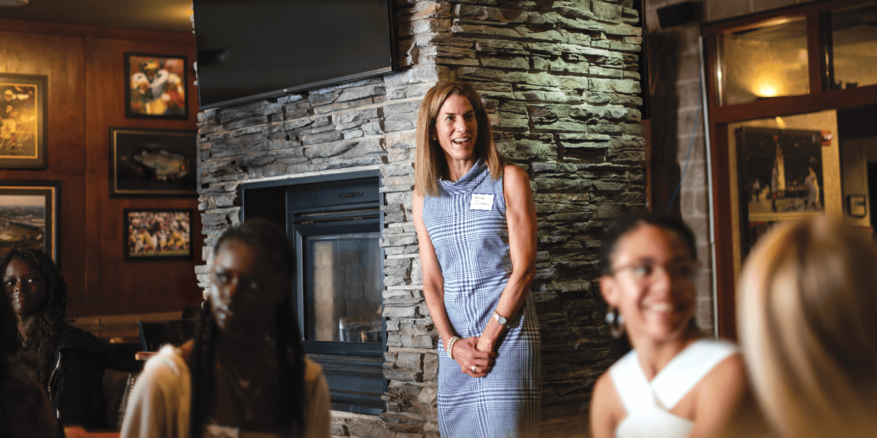 In this photo, Dean Anne Curzan stands at the center of a photo, smiling as she looks to her left, as she speaks to a group of students. In the foreground are four undergraduate students. The dean stands in front of a large fireplace.