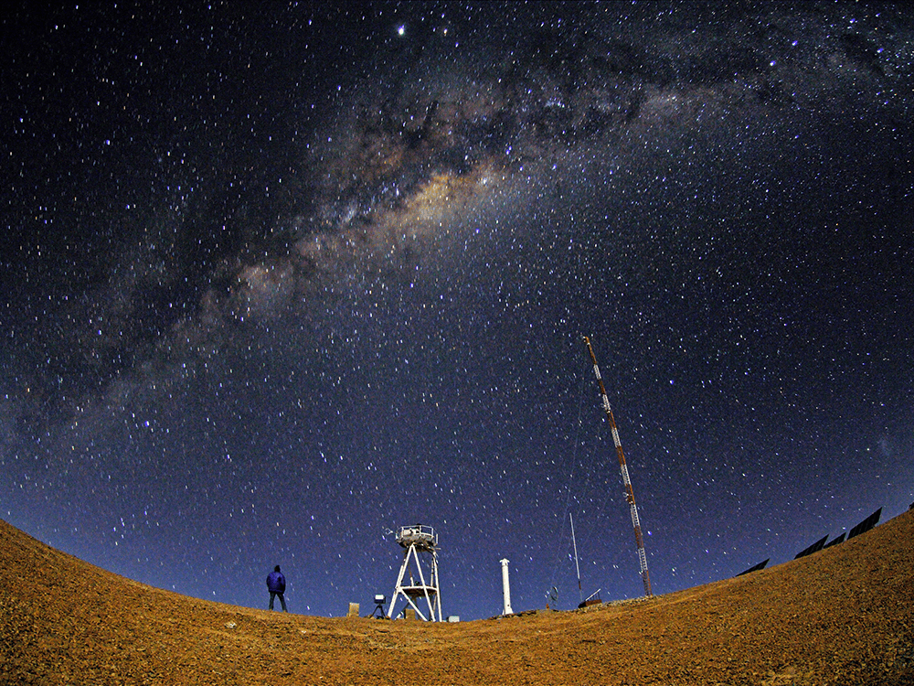 A night panoramic view of the Chilean Atacama Desert with small stars scattered across the sky. A person in a dark blue jacket and dark pants looks up at the sky where a clear band of the Milky Way galaxy is in the middle of the image. In front of the person in the middle ground is a white watchtower and other equipment.