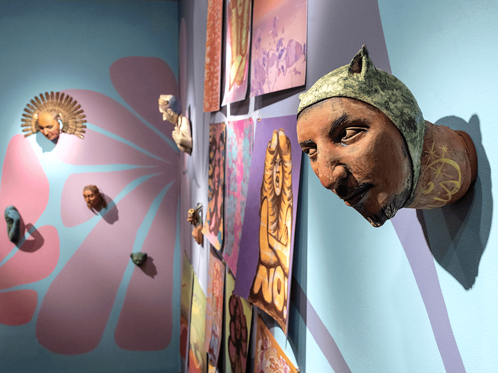 Brightly colored artwork is displayed on a wall that is light blue, pink, and purple. In the foreground is a three-dimensional ceramic piece that looks like a human male’s head coming out of the wall. In the background are paintings and more sculptural pieces. They are part of the exhibition “With Care” by artist Nicole Marroquin.