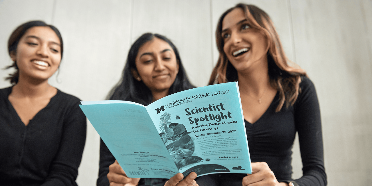Three female students sit together, all smiling and wearing black clothing, before a dance performance at the University of Michigan Museum of Natural History. They hold a blue pamphlet that says “Scientist Spotlight.”