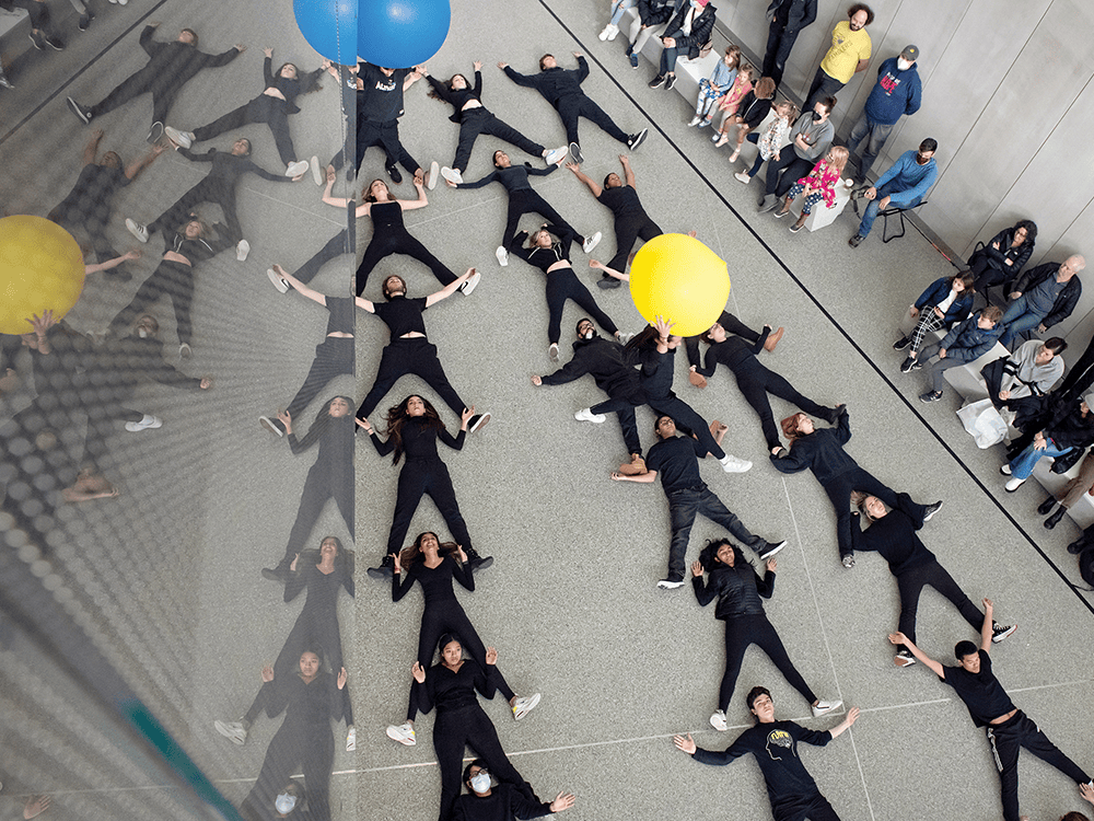 Twenty students wearing all black lie on the floor of the University of Michigan Museum of Natural History; two additional students stand, one holding a large yellow balloon and one holding a large blue balloon. The students are part of a dance performance that represents cell division and other aspects of human biology.