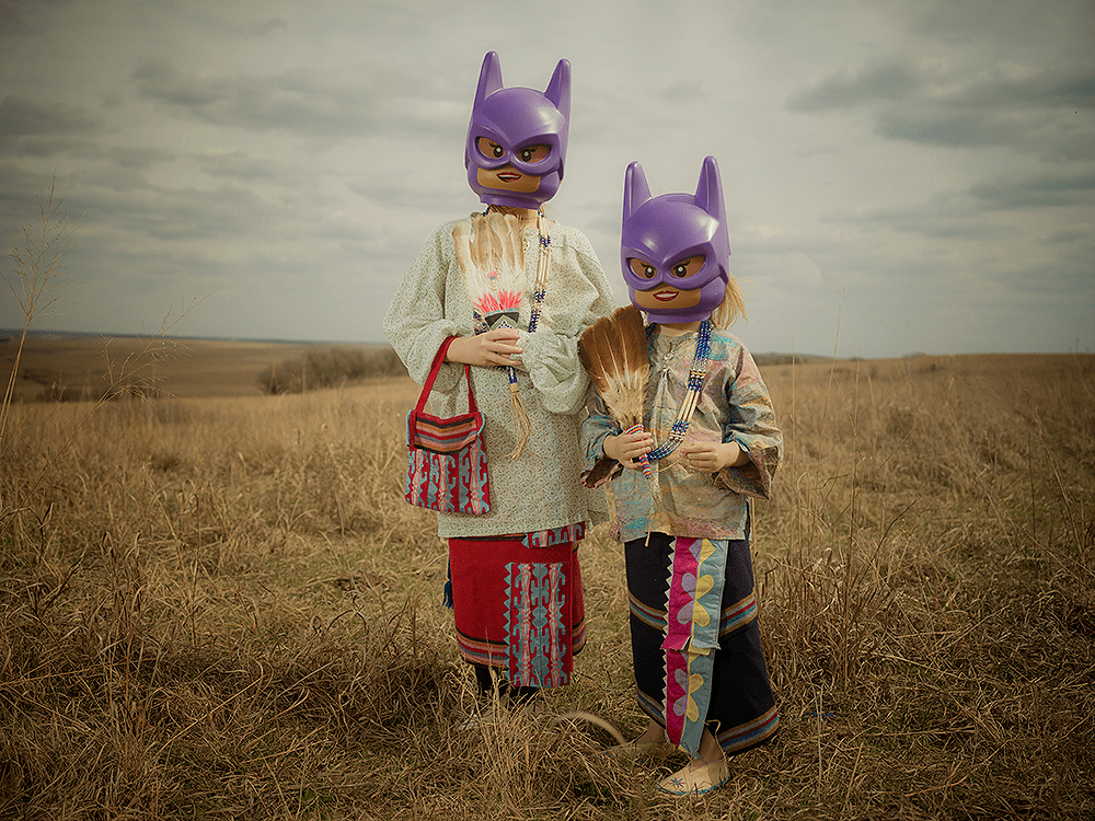Two Indigenous children standing in a field wearing Lego Batgirl Halloween masks
