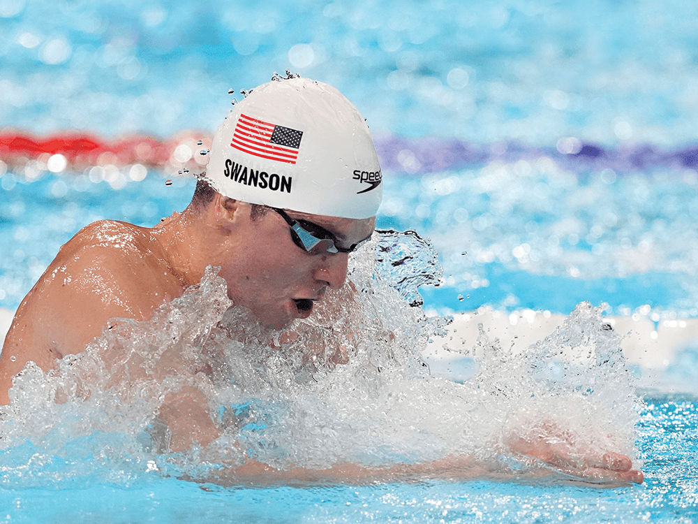 Charlie Swanson swims breaststroke in a pool, his mouth open as he takes a breath. He wears goggles and a white swim cap with an American flag.