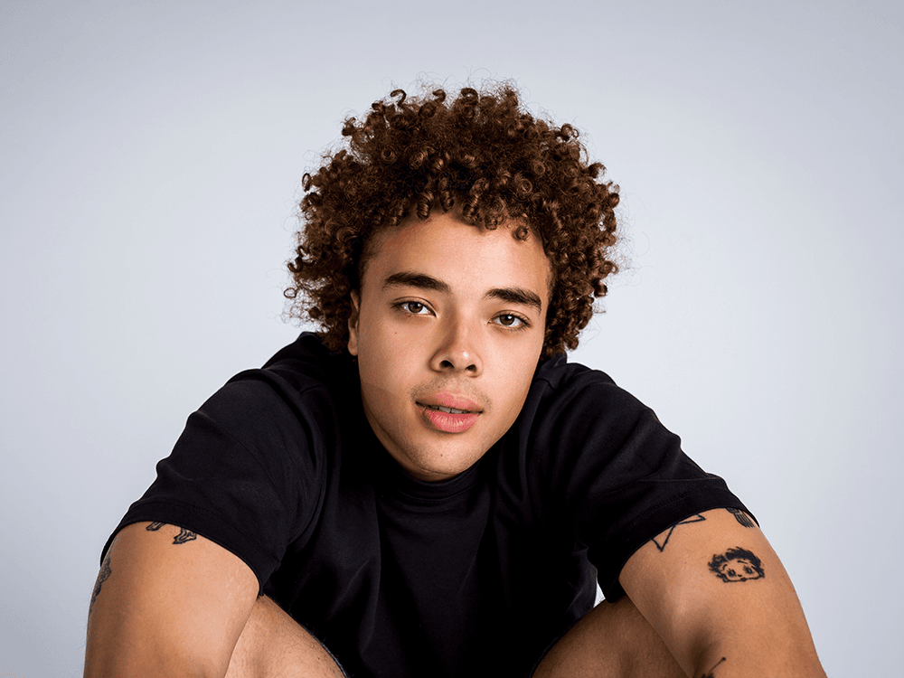 A photographic portrait of Daniel Williams, seated casually. Williams wears a black T-shirt and gazes directly at the camera. 