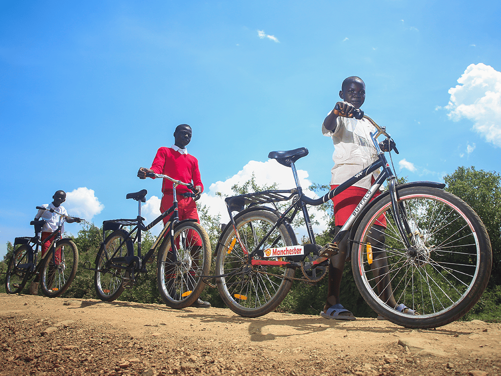 Three students stand in a row with their new bicycles on a dirt road.