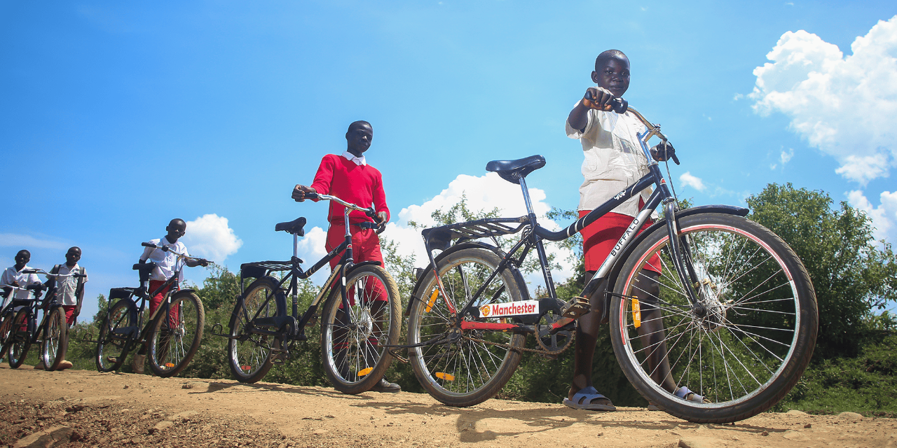 Three students stand in a row with their new bicycles on a dirt road.