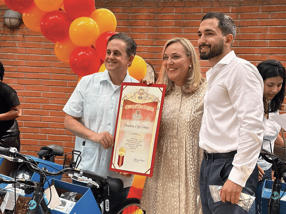 Three people pose for a photograph in front of a brick wall and balloons. Together, they hold a framed certificate.