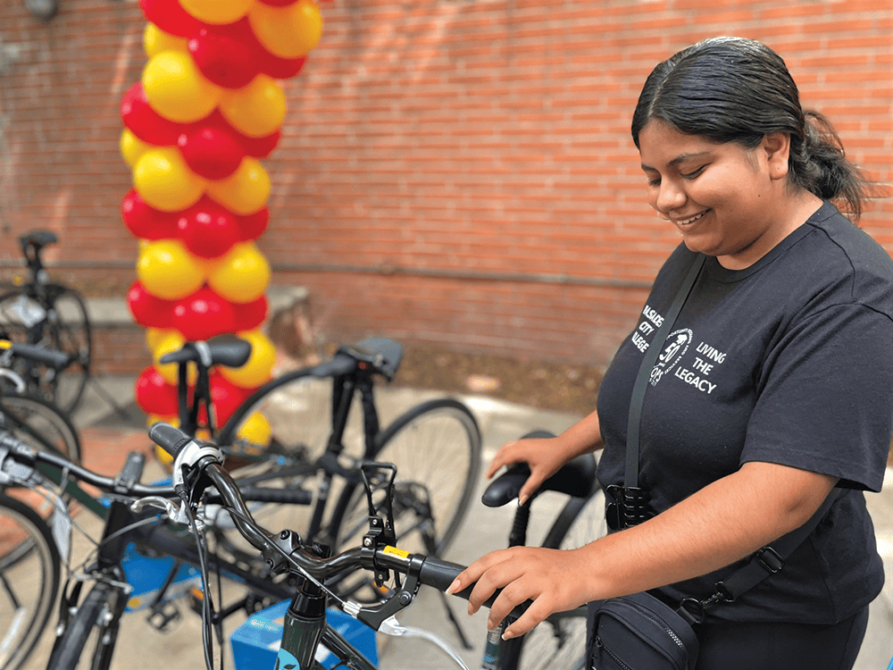 A student at Pasadena City College admires their new bicycle in front of a brick wall and colorful balloon arch.