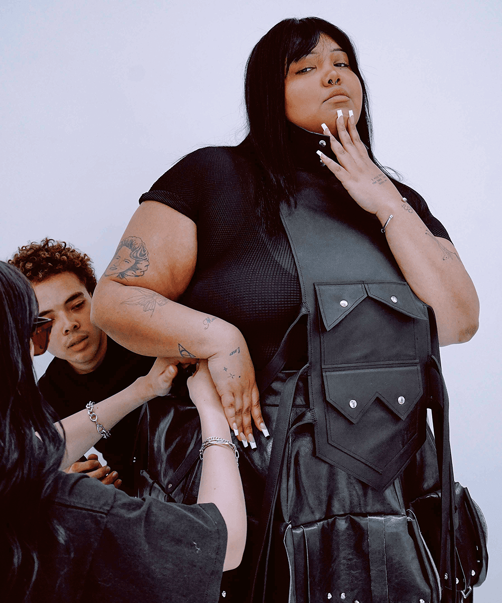 A person wearing an elaborate black leather pinafore stands in the foreground. Two people stand beside her, styling her clothing. 