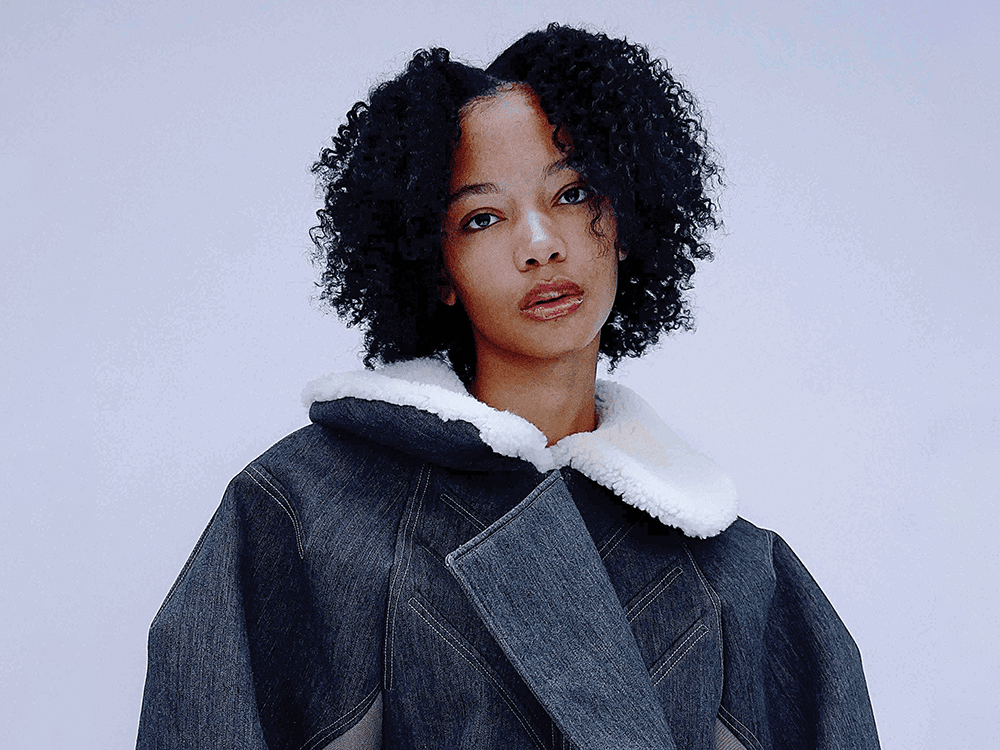 A person wearing an oversized denim jacket with a fur collar gazes directly at the camera.