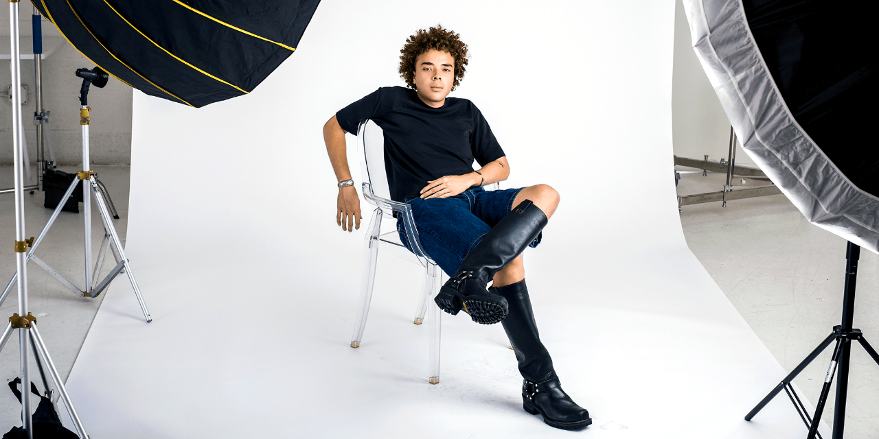 Daniel Williams sits in a clear plastic chair on a photography set, wearing a black T-shirt, denim shorts, and black motorcycle boots.