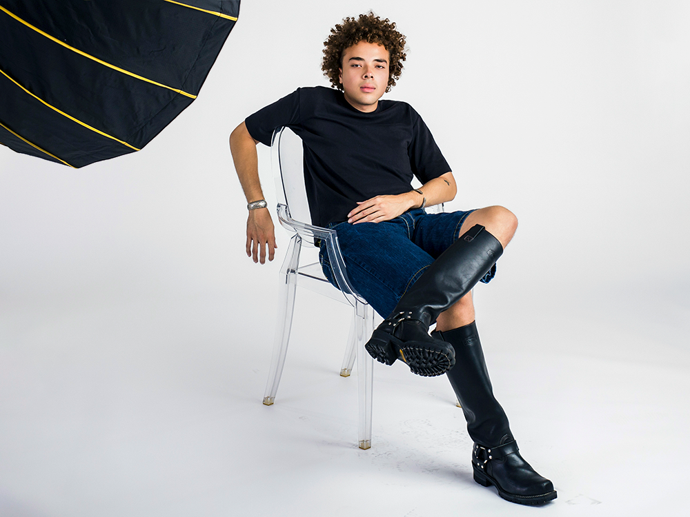 Daniel Williams sits in a clear plastic chair on a photography set, wearing a black T-shirt, denim shorts, and black motorcycle boots. 