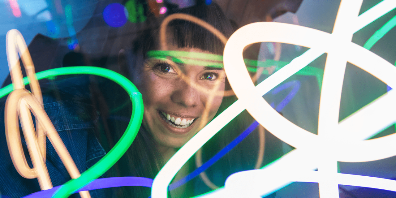 A photograph of Julianna Loera-Wiggins’s smiling face. She has dark hair and wears a denim jacket. Neon lights flash over her face. 
