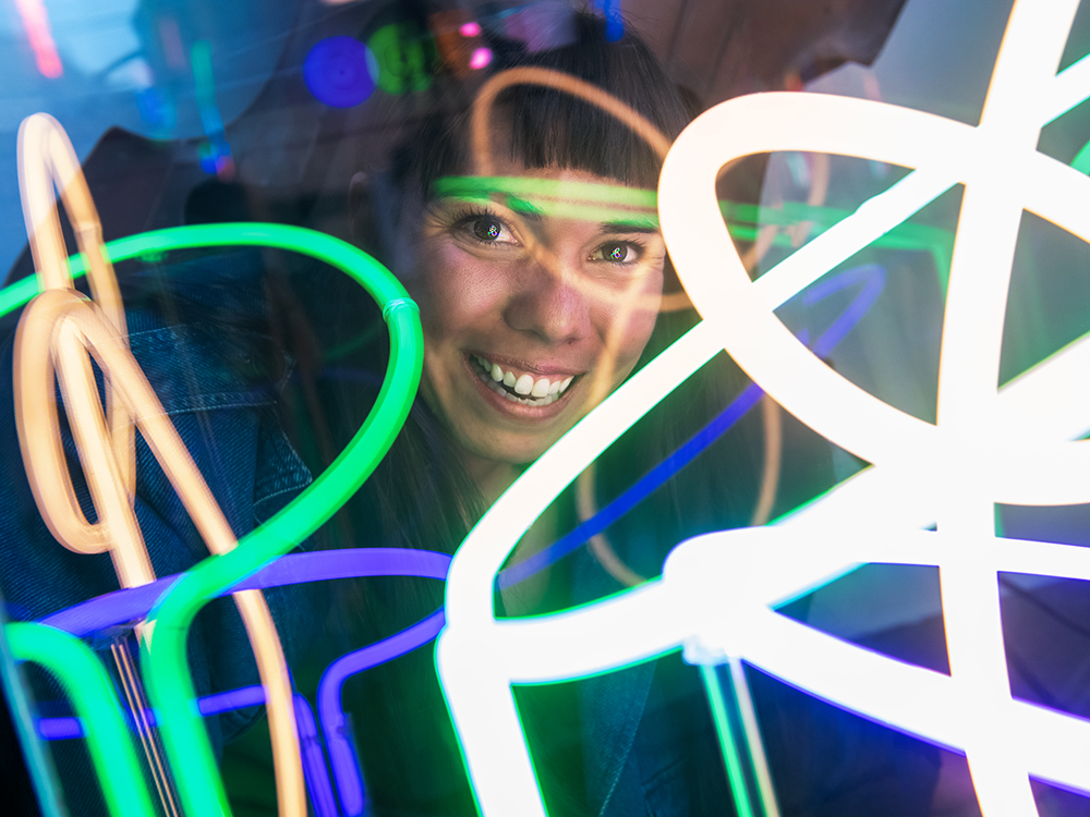 A photograph of Julianna Loera-Wiggins’s smiling face. She has dark hair and wears a denim jacket. Neon lights flash over her face. 