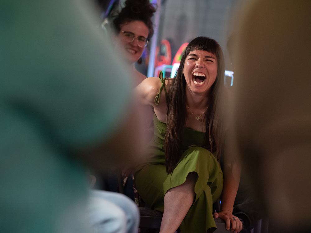 A photograph of Julianna Loera-Wiggins, seated and laughing joyfully with her mouth open. The foreground is blurred.