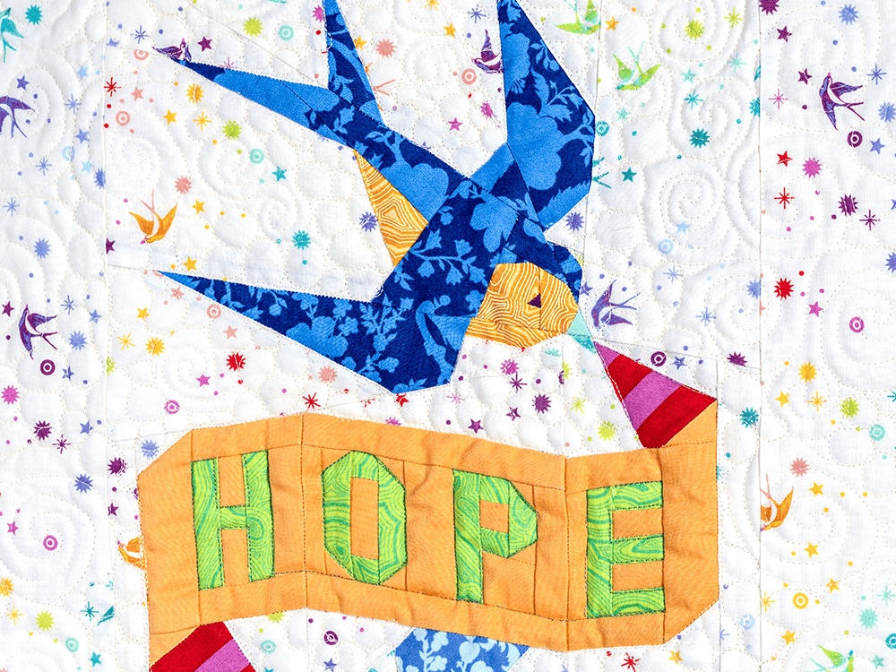 A section of a quilt includes a blue bird and a banner that says “hope.”