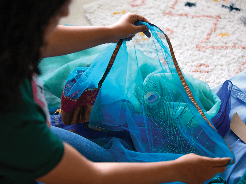 Supriya Kelkar’s arms frame this photograph. She holds diaphanous blue fabric trimmed in gold, a peacock feather, and two other blue fabrics.