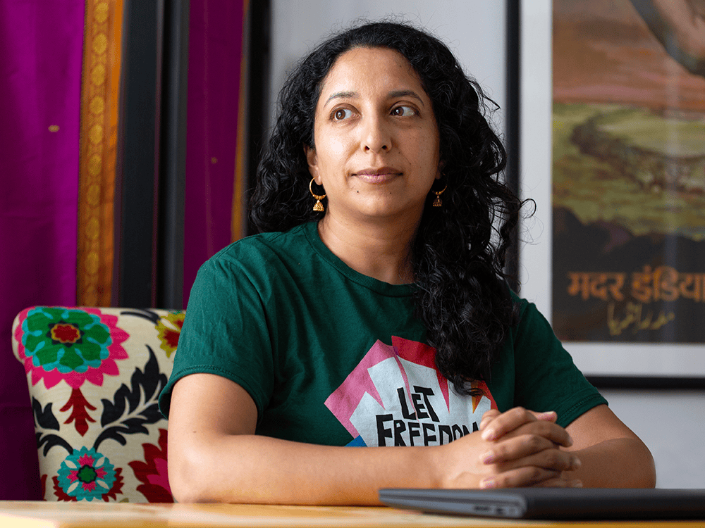 A photograph of Supriya Kelkar. Seated in a colorful chair, she has long dark hair and wears gold earrings and a green shirt.