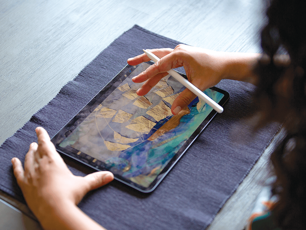 Supriya Kelkar’s hands hold a digital device and stylus. On the screen is a collaged illustration of a ship. 