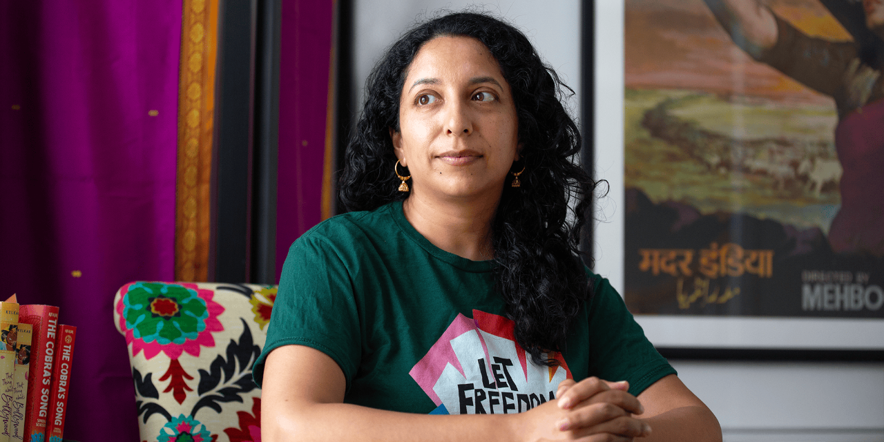 A photograph of Supriya Kelkar. Seated in a colorful chair, she has long dark hair and wears gold earrings and a green shirt.