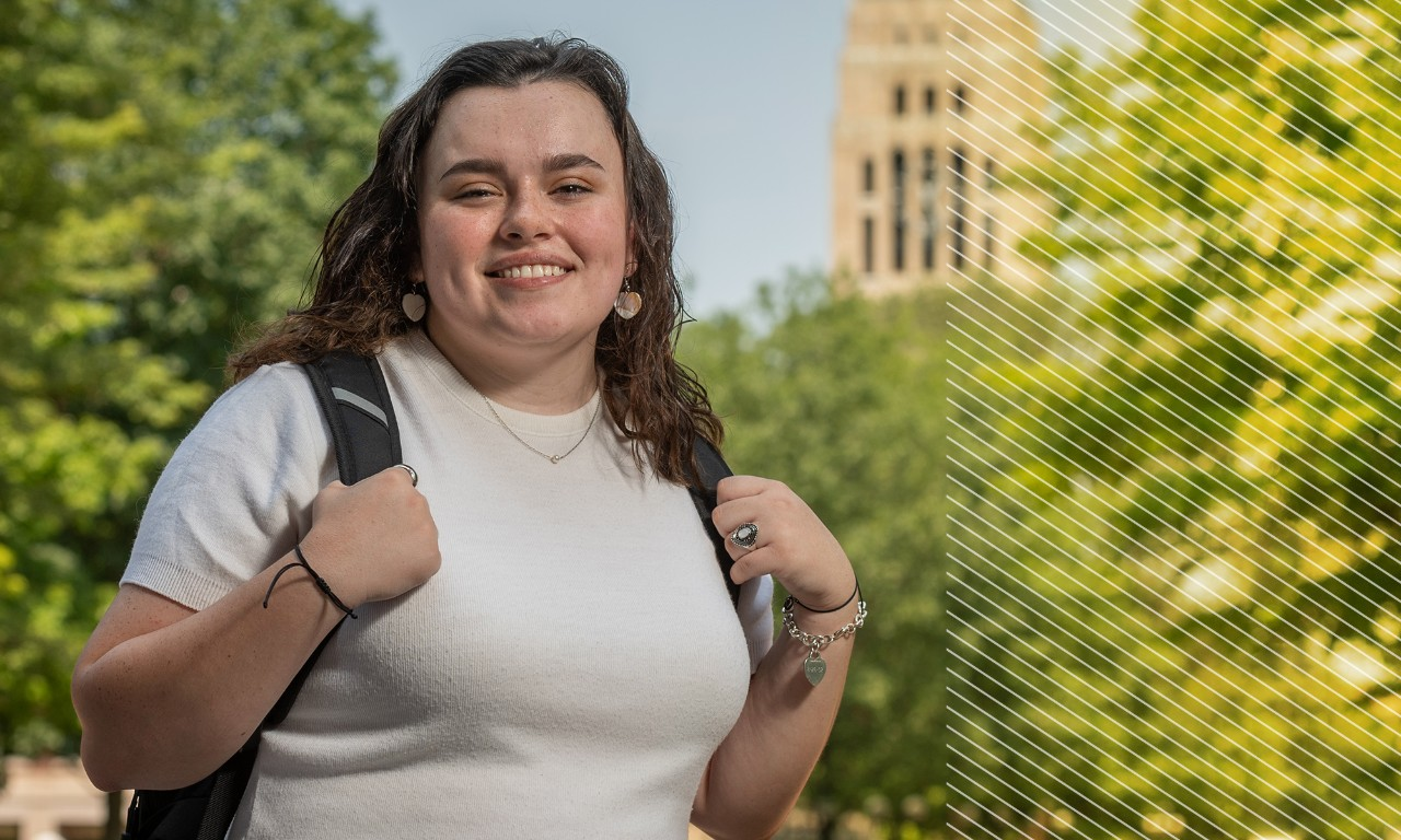 A student stands on the U-M Diag, with the clock tower in view behind her. She wears a white sweater and a backpack. 