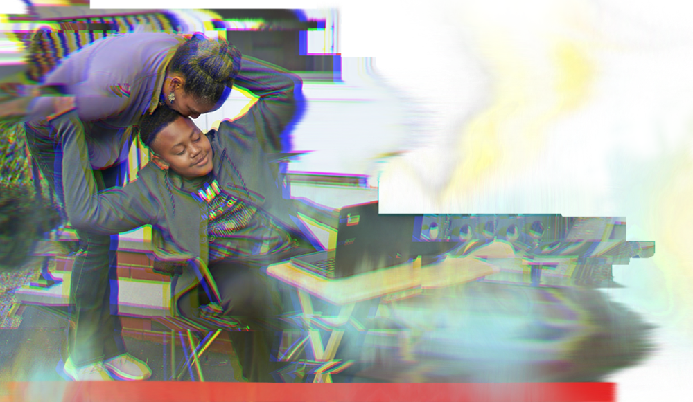 Image of a woman bending to kiss the head of a teenaged boy who is sitting at a table working on a laptop.