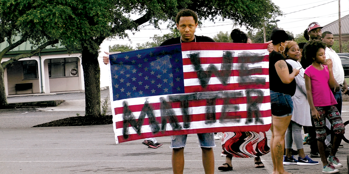 A movie still from a Brave New Films production in which a person of color holds an American flag painted with the words "We Matter."
