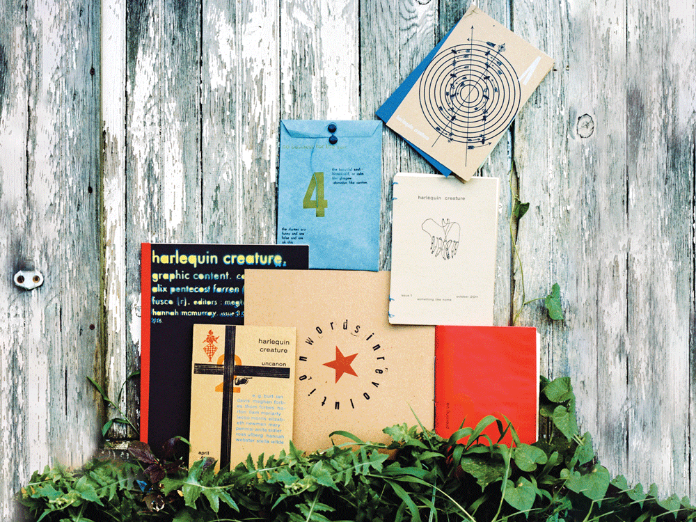 Several colorful, handbound issues of the harlequin creature literary journal are stacked against a wooden fence. 