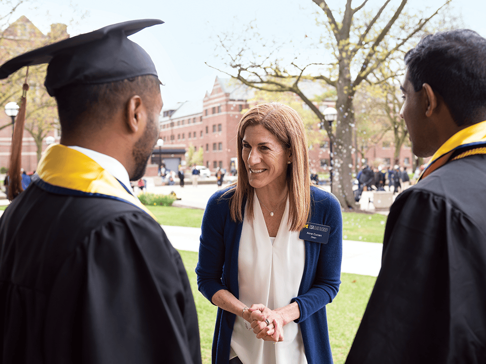 In a photo, Dean Anne Curzan speaks with two men wearing graduation gowns. She is smiling and facing the camera. The graduates’ backs face the camera.