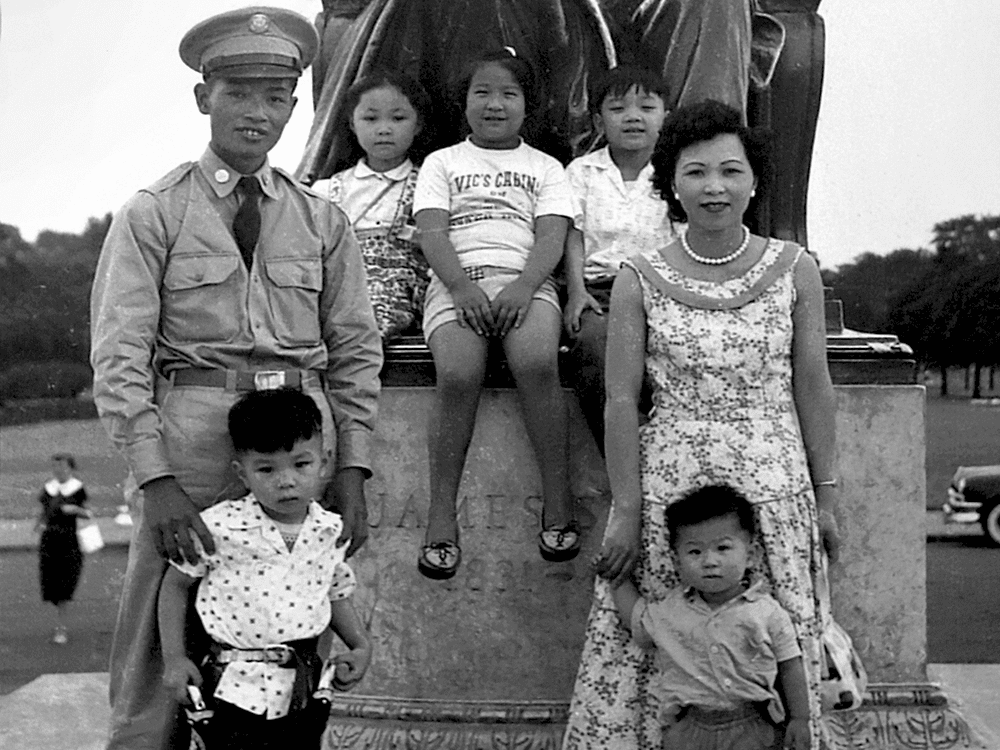 A black and white photo circa 1956 of a family in front of a statue in a park. There are five children and two adults.