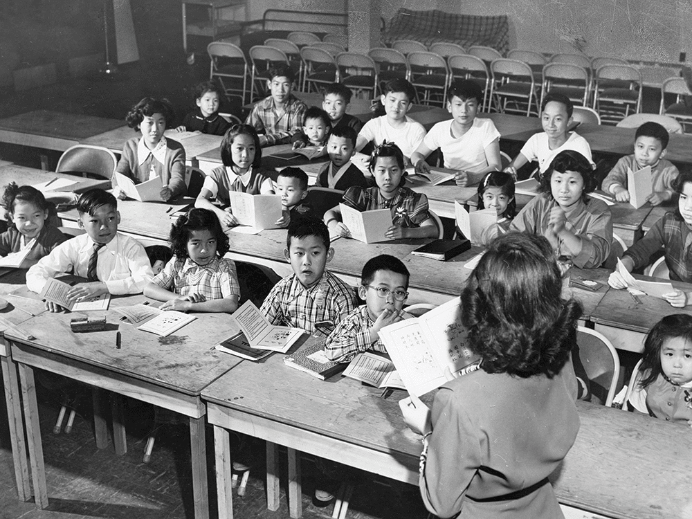 A black and white photo circa 1950s of a classroom. Several children sit in three rows of desks and the back of a teacher’s head is visible. 
