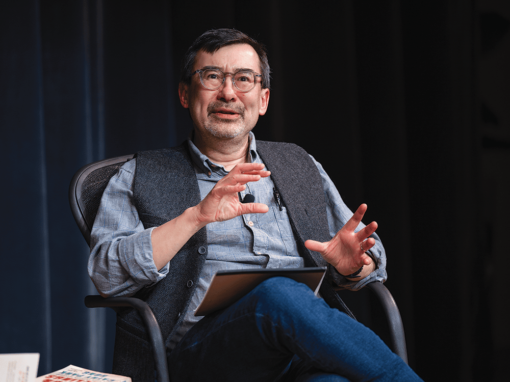 A photo of Peter Ho Davies, a man wearing glasses and a vest, seated in a chair and speaking. 
