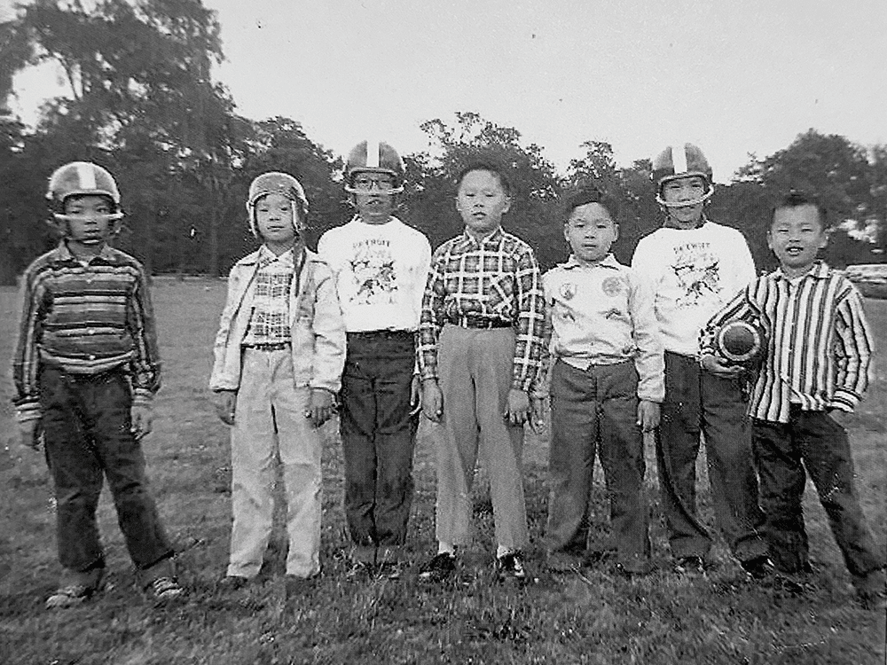 A black and white photograph of a group of seven Chinese American children, four of whom wear football helmets, and one of whom holds a football, smiling at the camera.
