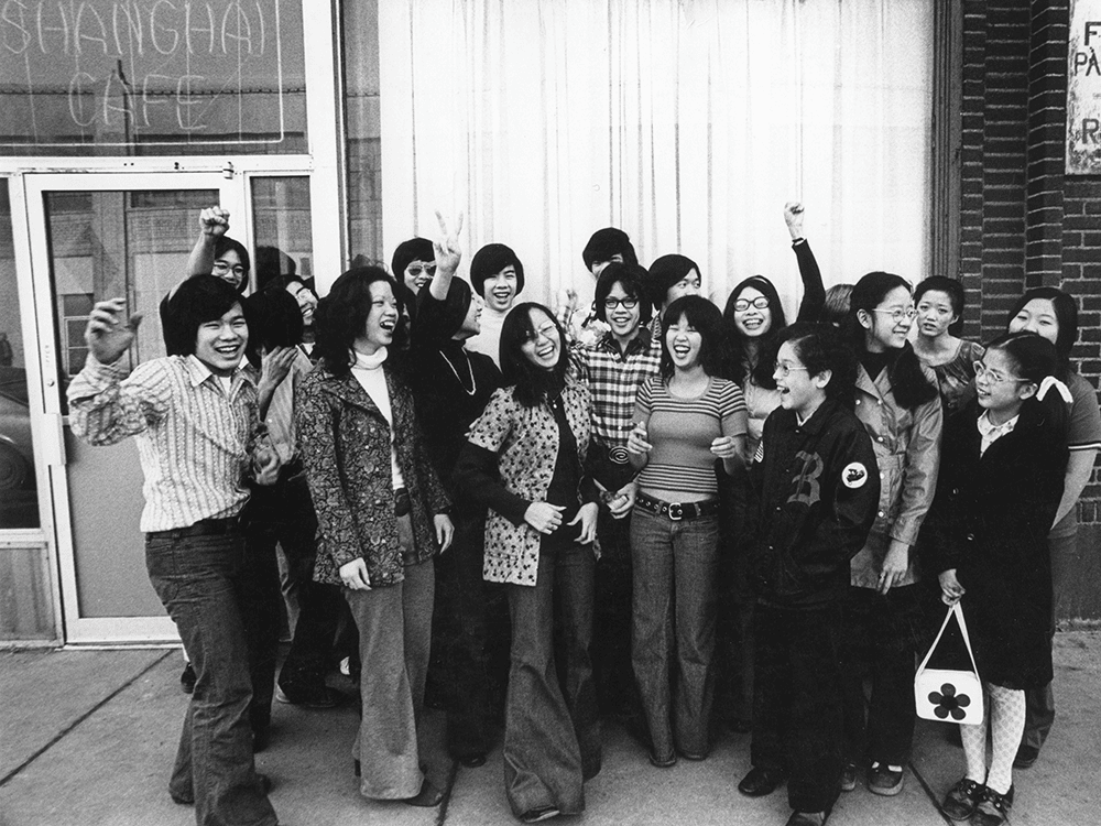 A black and white photo of a group of Chinese American youth gathered together and smiling joyfully in 1970s clothing.