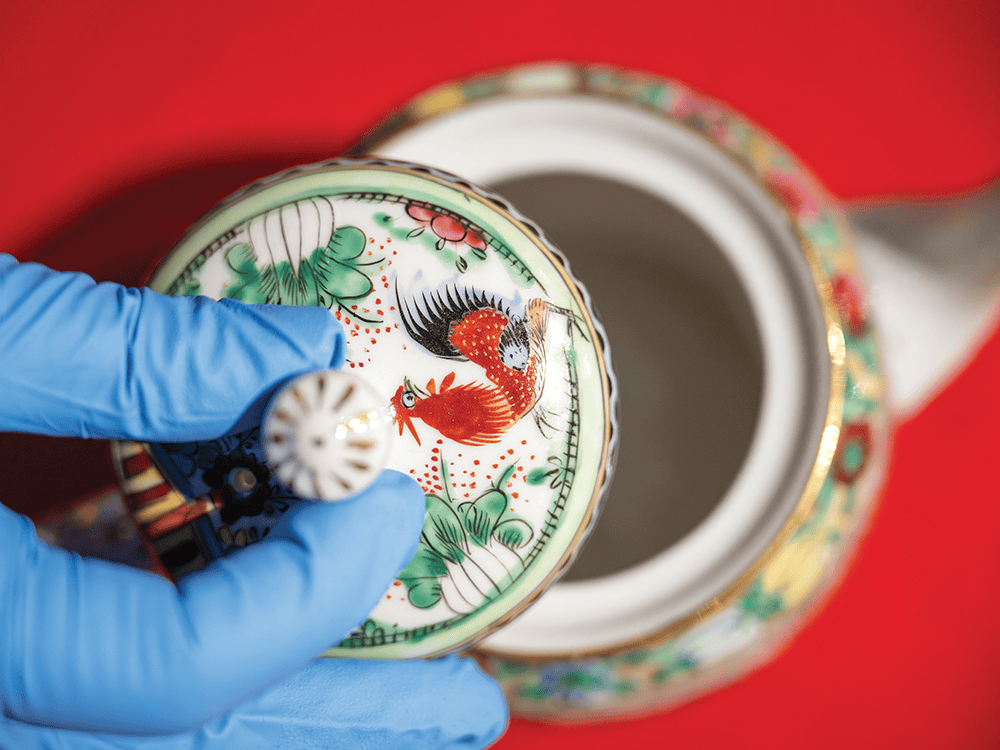 A photo of a blue vinyl-gloved hand lifting the top off of an ornately painted teapot. 