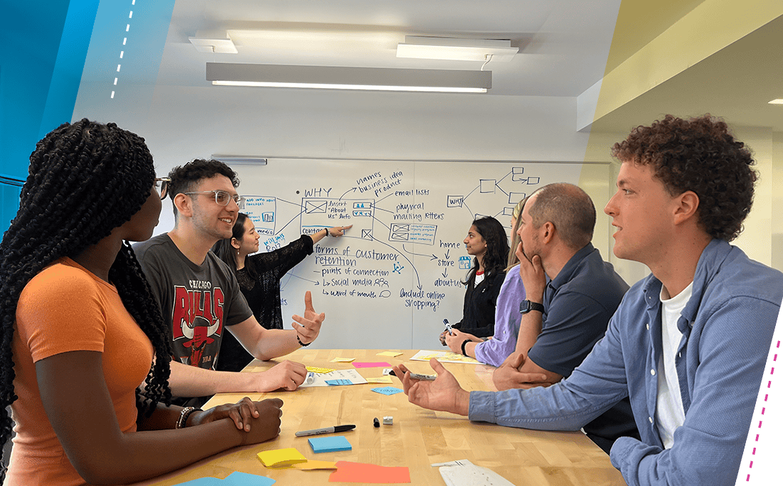 Students seated at a table converse with one another while students in the background work together on a whiteboard. 