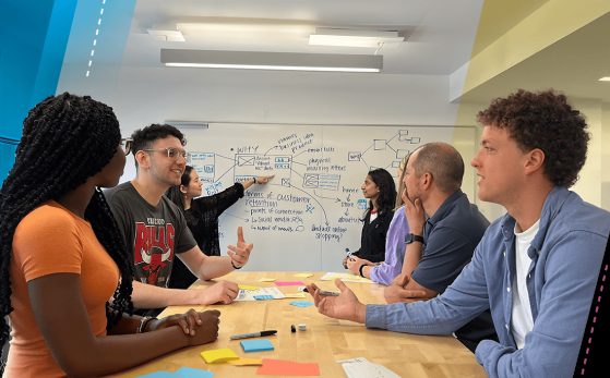 Students seated at a table converse with one another while students in the background work together on a whiteboard. 