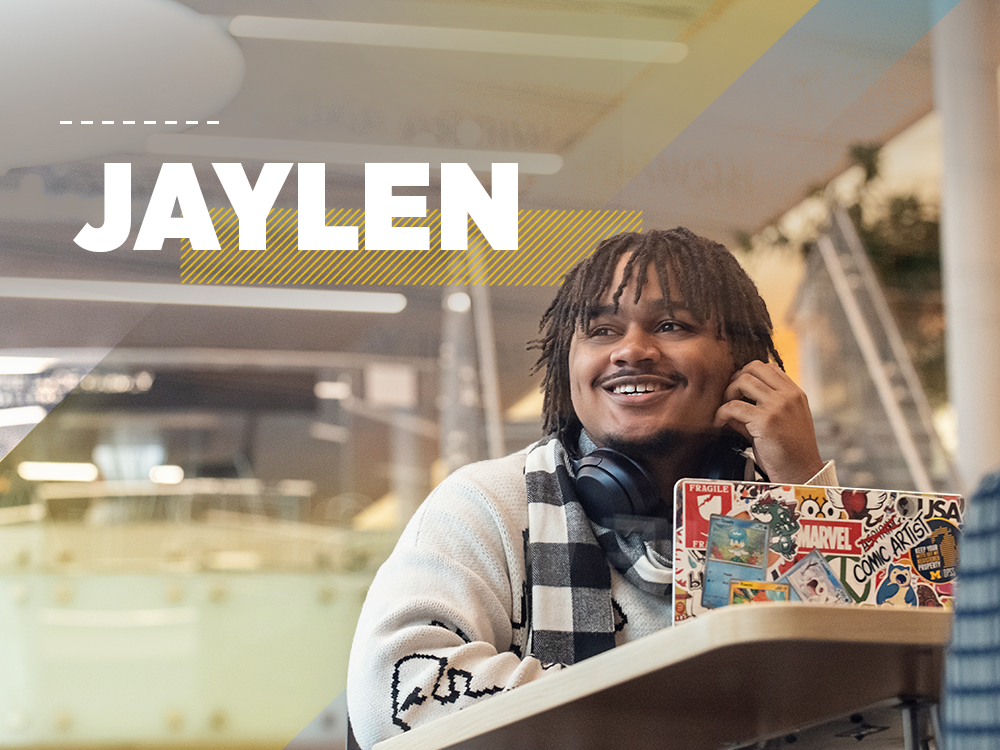 Jaylen is sitting at a desk with a laptop covered in stickers, he is smiling and looking away from the camera. Text on the image reads "Jaylen".