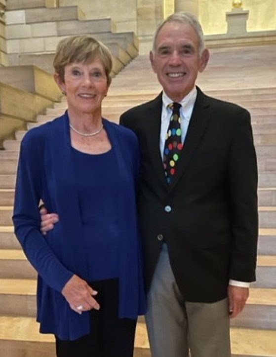 A man and woman stand smiling in front of a marble staircase. They are in their early to mid 70s. The woman wears a dressy royal blue top and matching drapey open front sweater with black slacks and high-heeled sandals. The man wears a white button down shirt, dark suit jacket and a tie with multi-color dots against a dark background, with tan slacks and black dress shoes.