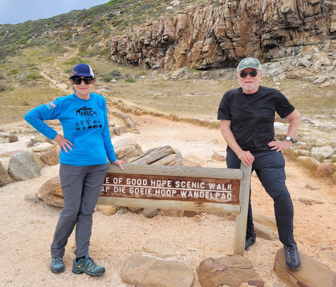 Sidonie (Sid) Smith and William (Bill) Kinley are pictured on a trip to South Africa. They pose on a hiking trail, each standing at either side of a sign that says "Cape of Good Hope Scenic Walk" in English and Afrikaans. Sid wears a cap, a blue long-sleeved shirt, gray pants, and hiking boots. Bill wears a black short-sleeved shirt, gray pants, and hiking boots.