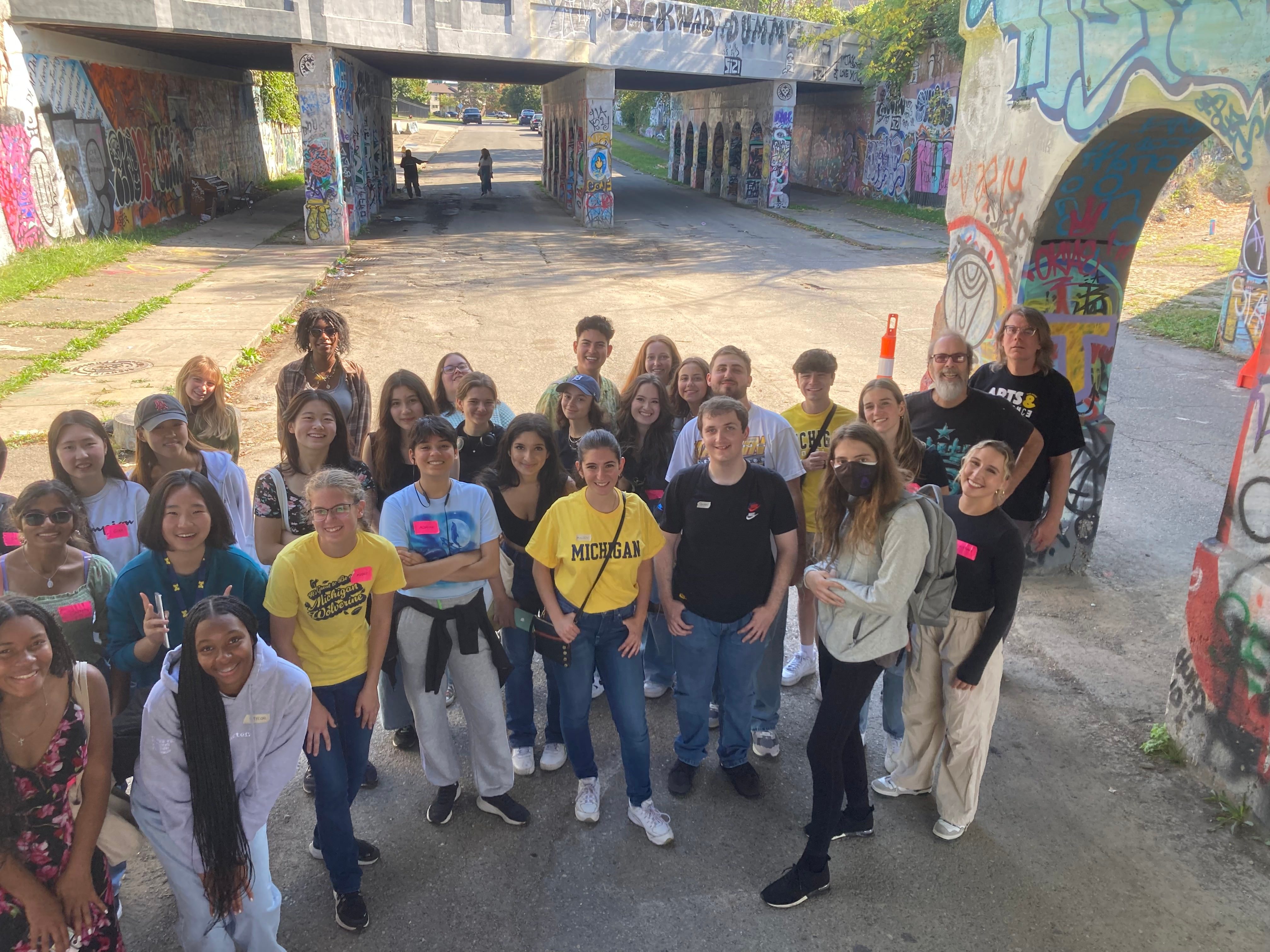 A diverse group of about 30 LSWA students pose in front a colorful, graffiti-covered highway underpass in Detroit. They are smiling.