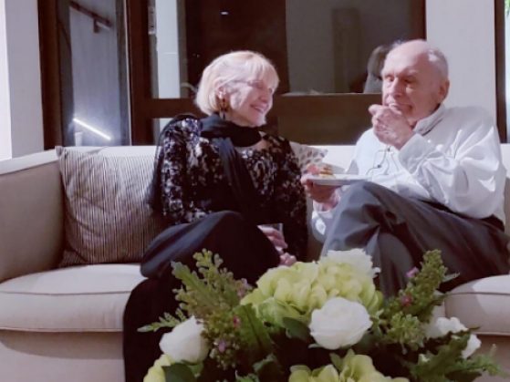 Dr. Priscilla S. Rogers and Dr. Gunter Dufey pictured engaged in a conversation while smiling and seated together on a couch.