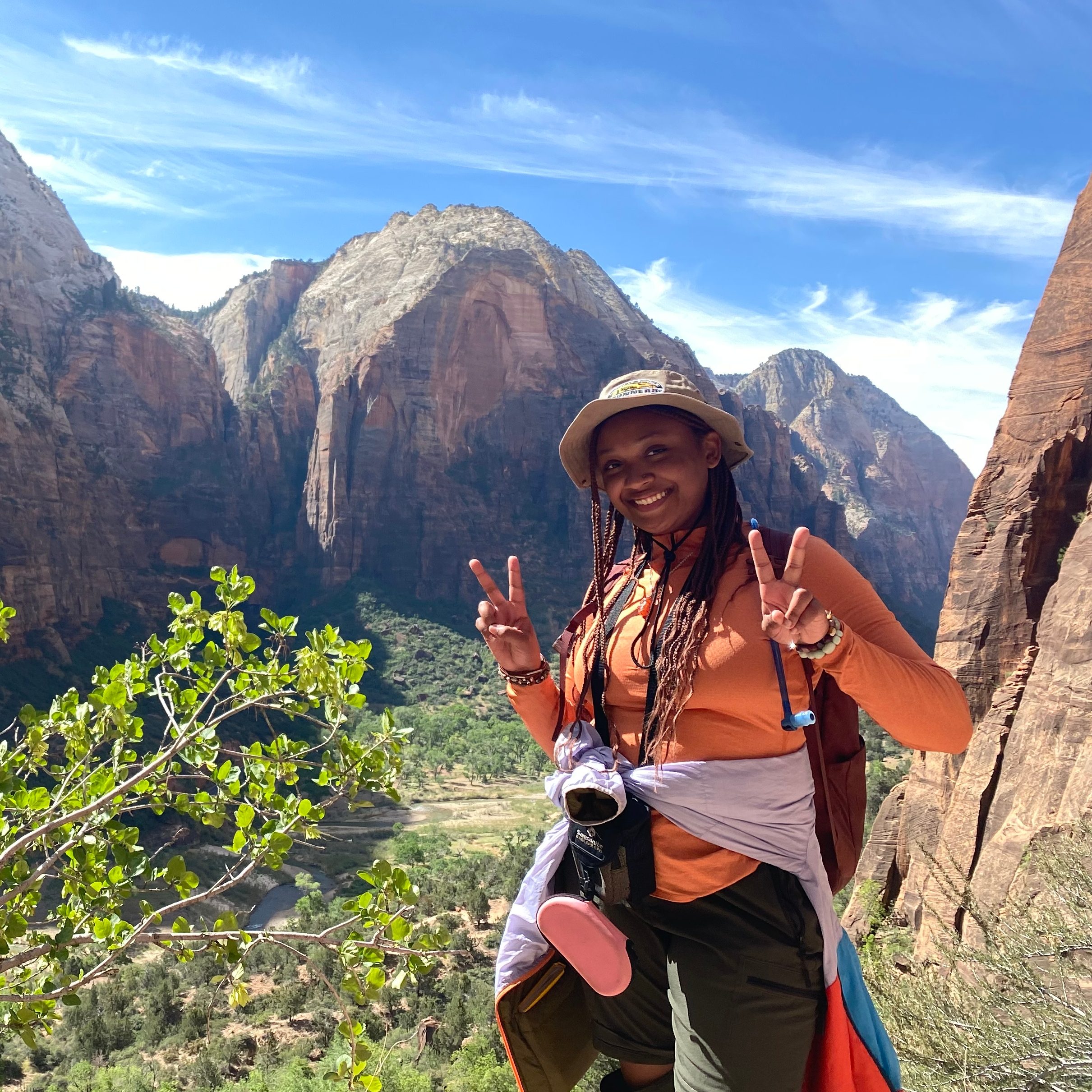 A female student flashes peace signs with her hands in front a mountain. The sky is blue, sun is shining. The student is African American, with a khaki bucket hat over long dark braids; she is wearing a khaki bucket hat, orange long sleeve shirt and olive green trek pants. She has a jacket tied around her waist.