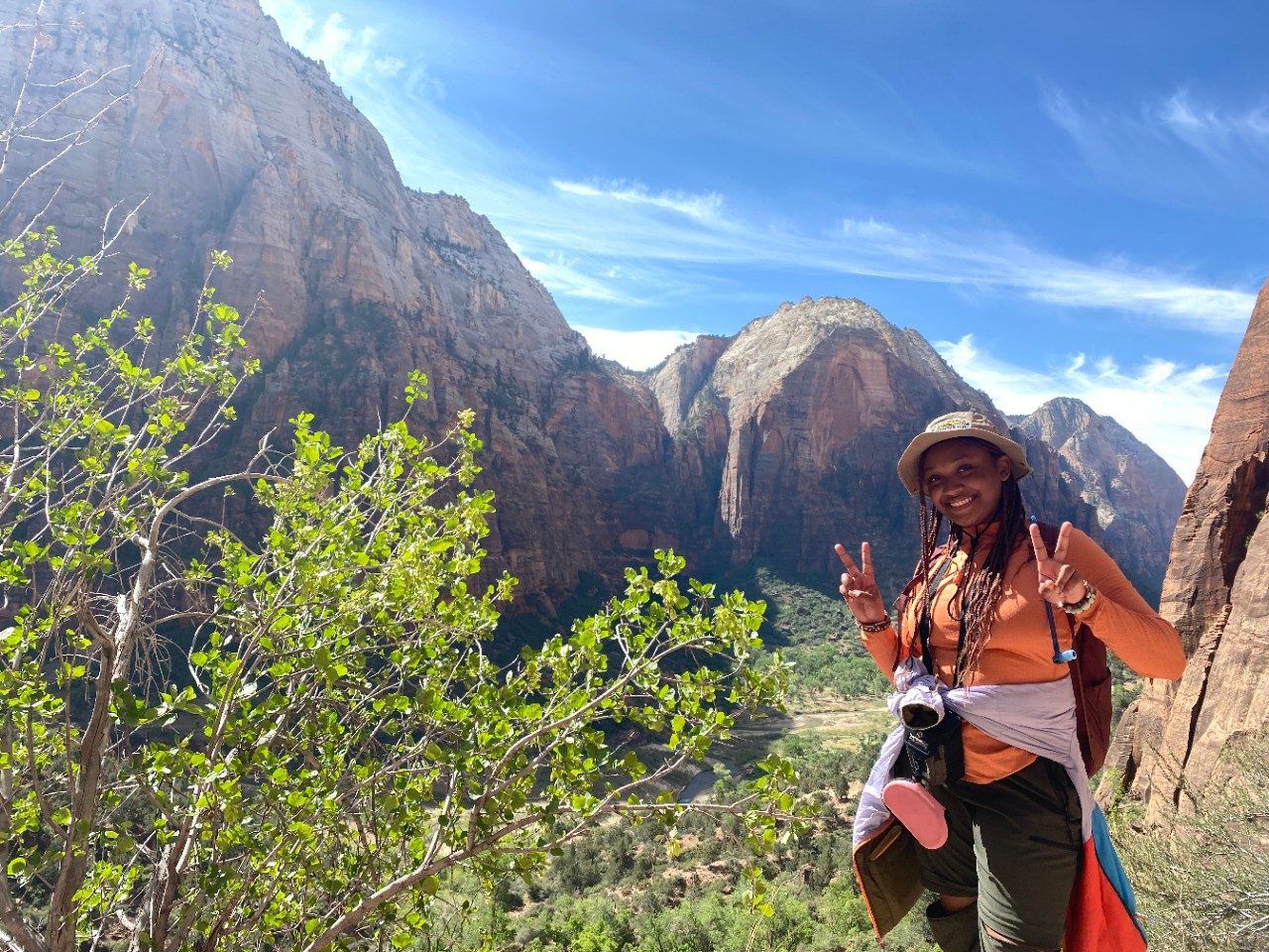 A female student flashes peace signs with her hands in front a mountain. The sky is blue, sun is shining. The student is African American, with a khaki bucket hat over long dark braids; she is wearing a khaki bucket hat, orange long sleeve shirt and olive green trek pants. She has a jacket tied around her waist.