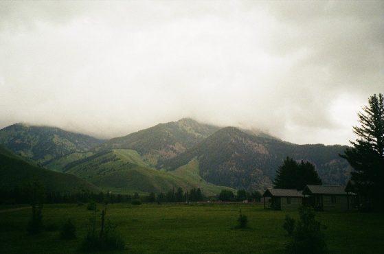 Small, rustic cabins sit in a green field at the base of a mountain shrouded in mist at Camp Davis Rocky Mountain Field Station in Jackson, Wyoming.