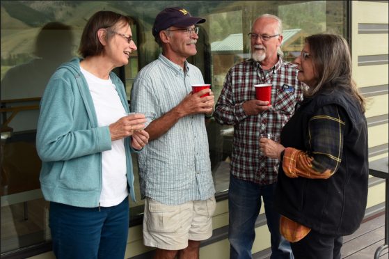 Four casually dressed alumni stand on the porth of a Camp Davis building holding red solo cups and clear plastic drink cups. Two of them are man with gray facial hair, one wears shorts, a short sleeved button down shirt and cargo shorts, and a navy blue baseball cap with a maize block M; the other wears a long sleeve plaid button down shirt and jeans. One woman, with a short brown bob wears jeans and turqoise zip sweatshirt over a white t-shirt; the other, with long brown hair, wears jeans and a dark vest over a plaid long sleeve shirt. All four wear glasses. They appear to be chatting; they are smiling.