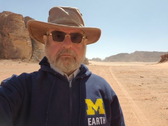 A man wearing a wide brimmed tan sunhat, button down shirt, sunglasses and navy blue full zip U-M EARTH  hoodie sweatshirt poses in the desert. He has a grayish mustache and close cropped beard.