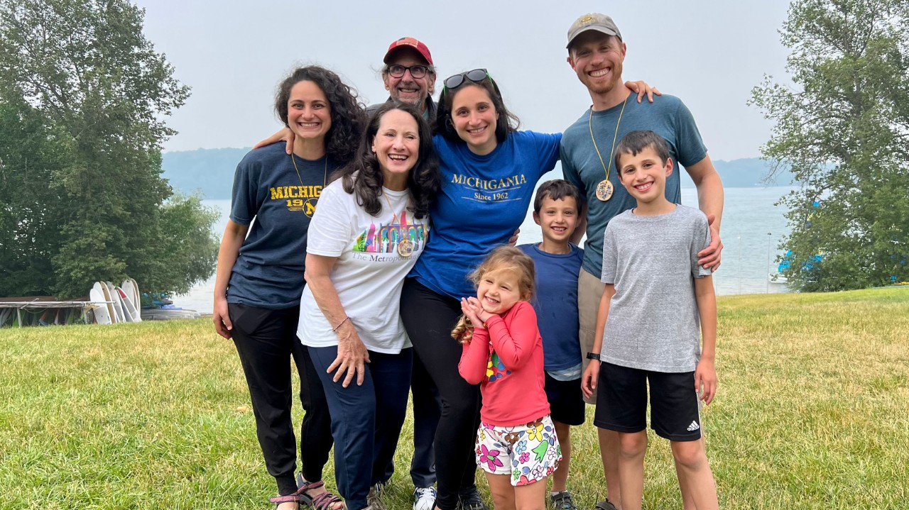 Eight people, including three young kids, two grandparents and three adults in their thirites, representing three generations of the Woll family, pose outside on the grassy lakefront at Camp Michigania. They are casually dressed in shorts and t-shrits and leggings, some are wearing ball camps. They are all smiling happily.